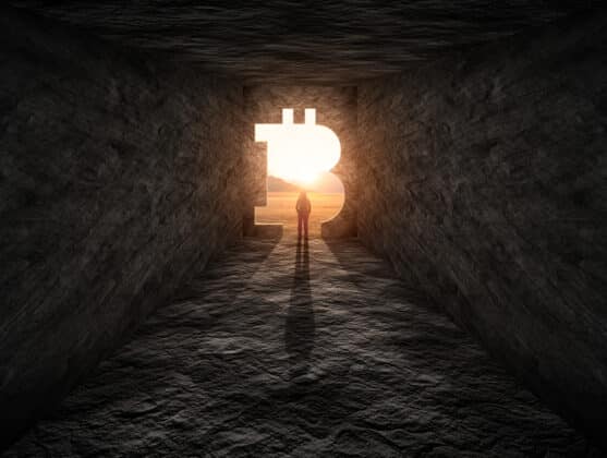 Photo of man standing over a cave entrance in a shape of Bitcoin. Photo of man standing over a cave entrance in a shape of Bitcoin.