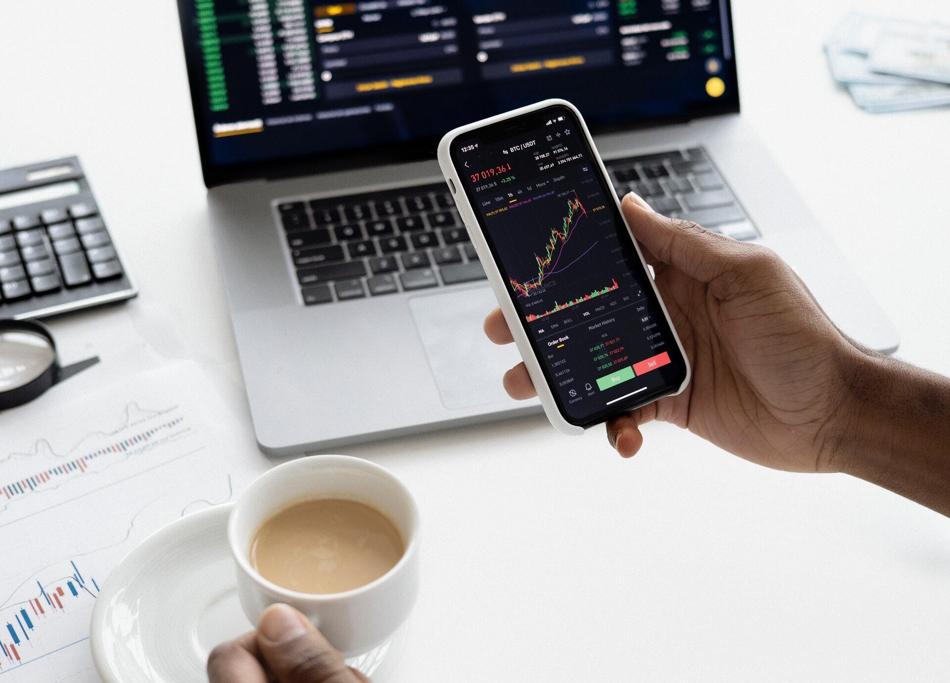Photo of a hand holding a smarphone displaying a candlestick chart on the screen.