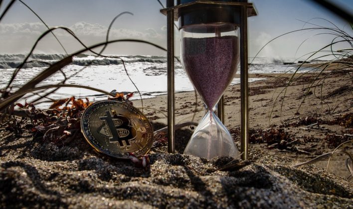cryptocurrency-3401786_1280 Photo of a bitcoin burried in sand with an hour glass next to it.